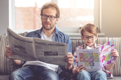 Father and son reading newspaper and book at home on a couch.