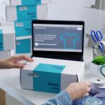 Person packing a "thank you" box near a laptop with a t-shirt webpage, surrounded by other boxes, ribbon, tape, a cup, plants, a phone, and a calculator on a bright desk.