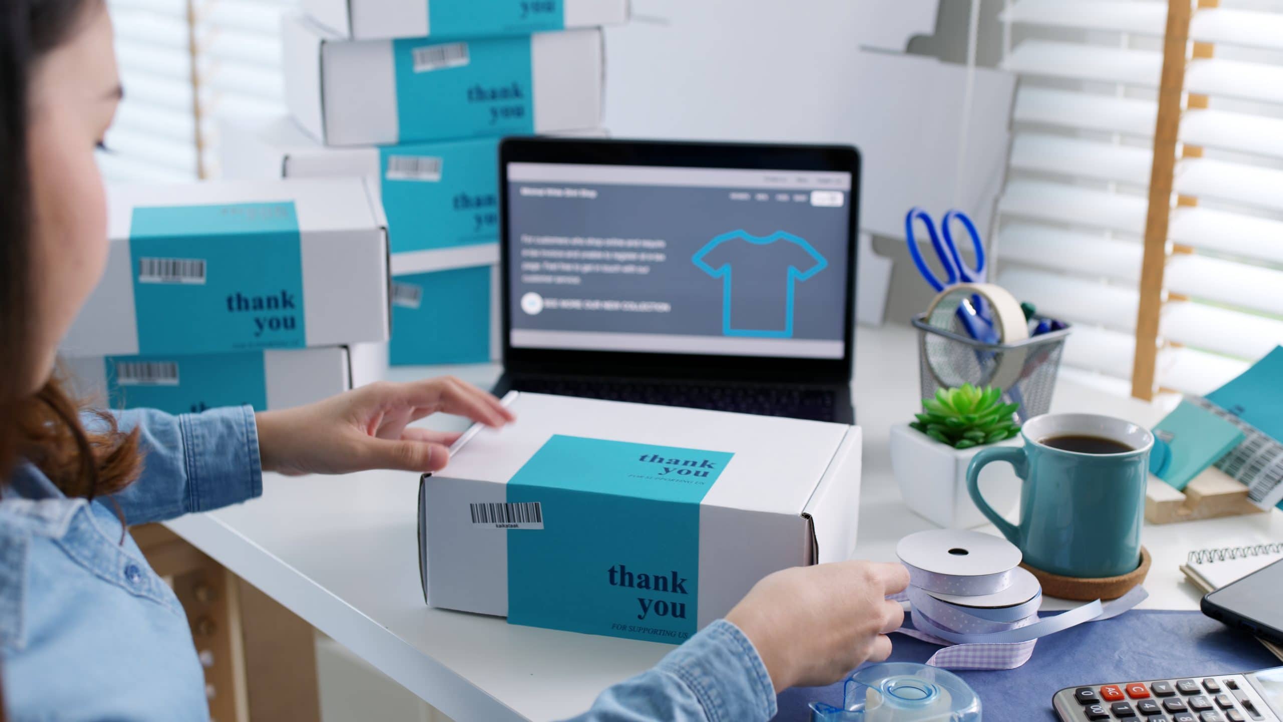 Person packing a "thank you" box near a laptop with a t-shirt webpage, surrounded by other boxes, ribbon, tape, a cup, plants, a phone, and a calculator on a bright desk.